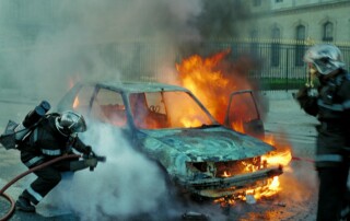 Firefighters extinguish a blazing car on a street, smoke rising around the vehicle.