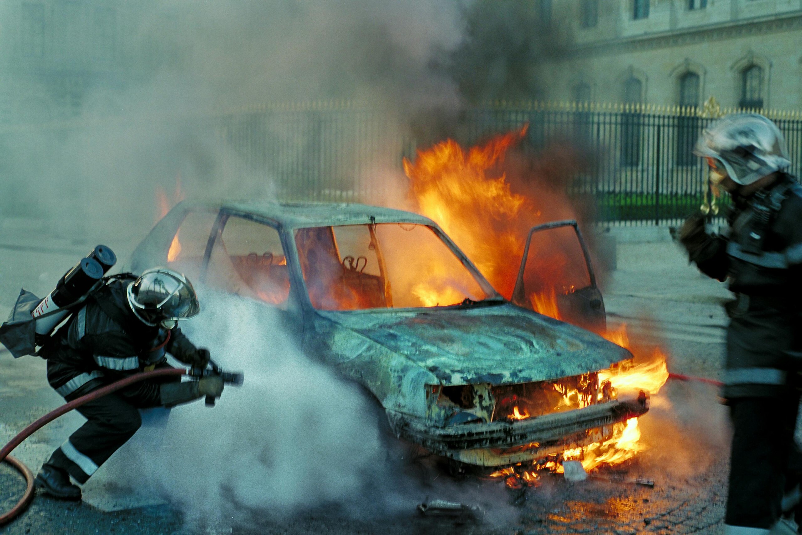 Firefighters extinguish a blazing car on a street, smoke rising around the vehicle.