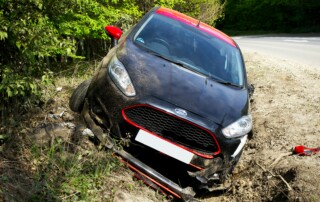 Black car in a ditch off a dirt road, front end damaged, daytime outdoors.