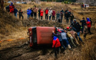 A red vehicle on its side in a muddy ditch as bystanders and rescuers watch and assist.