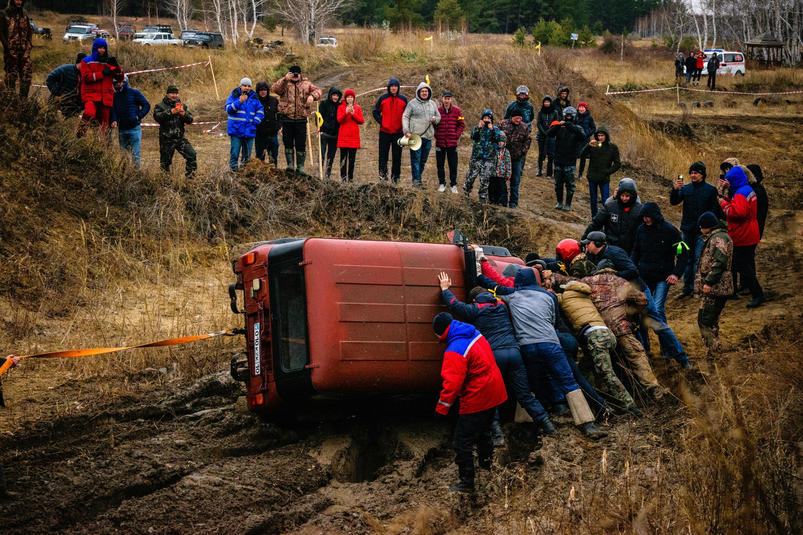 A red vehicle on its side in a muddy ditch as bystanders and rescuers watch and assist.