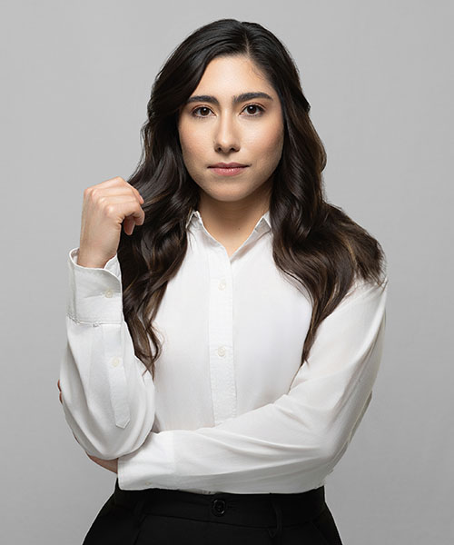 A woman in a white blouse and black trousers stands against a gray studio backdrop.