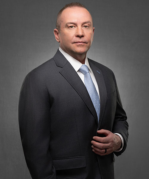 Portrait of a serious man in a dark suit and light blue tie against a neutral grey backdrop.
