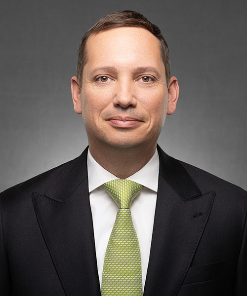 A man in a dark suit and lime-green tie smiles against a gray studio background.