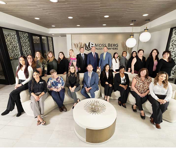 Group photo in a modern office lobby; people stand behind a circular coffee table, with Moss Berg Injury Lawyers sign in the background.