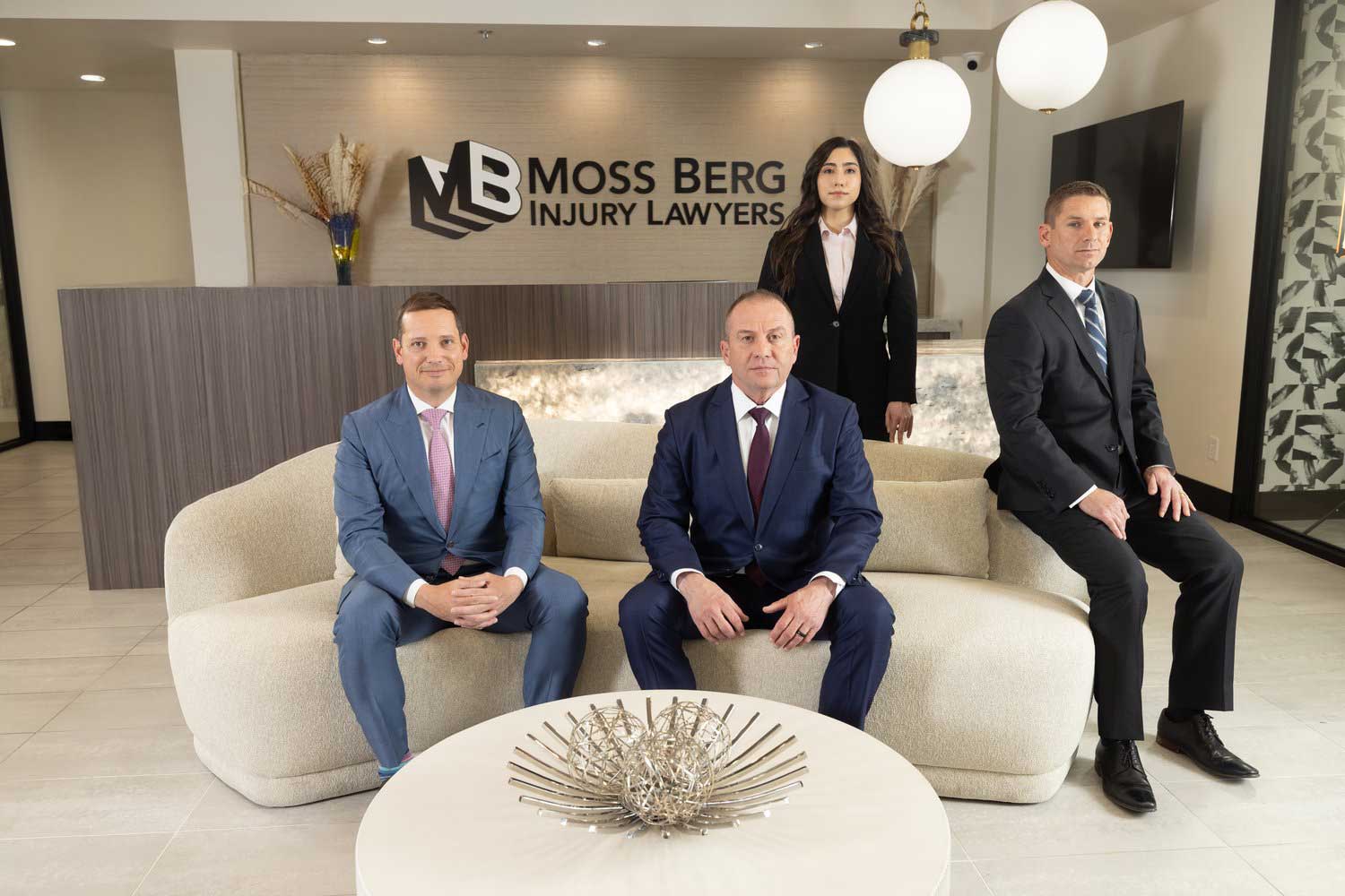 Lawyers pose in a modern office lobby; two seated on a sofa, two standing behind, Moss Berg Injury Lawyers sign.
